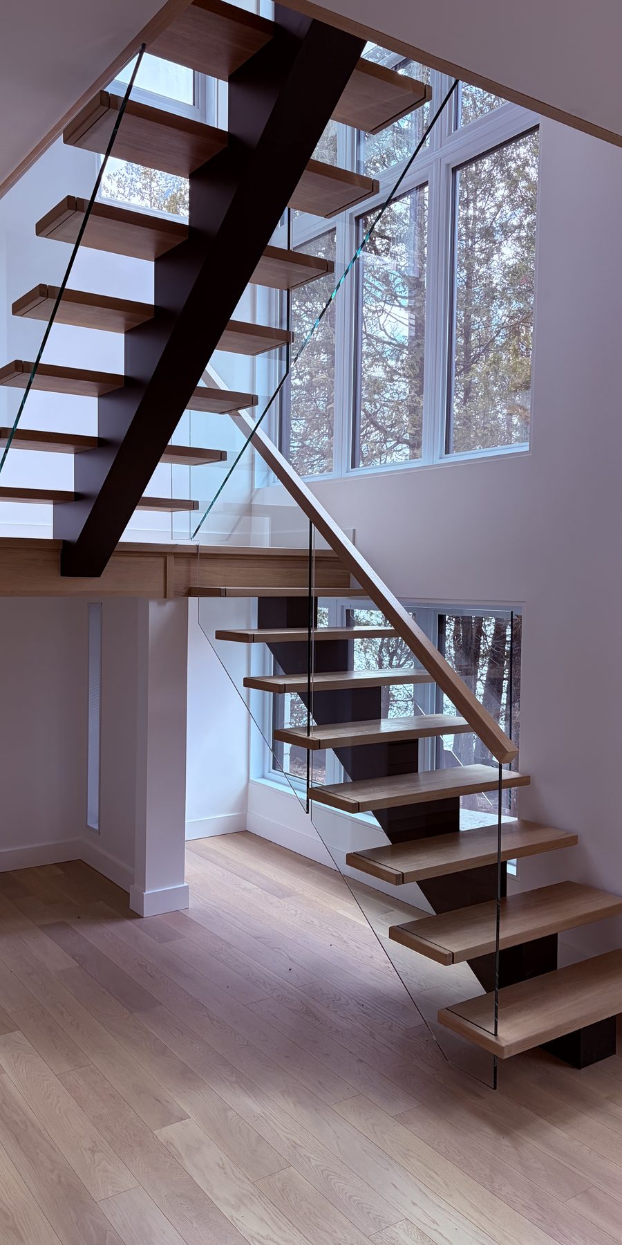 Escalier suspendu à limon central d'acier, marches de chêne et garde-corps de verre devant une baie vitrée